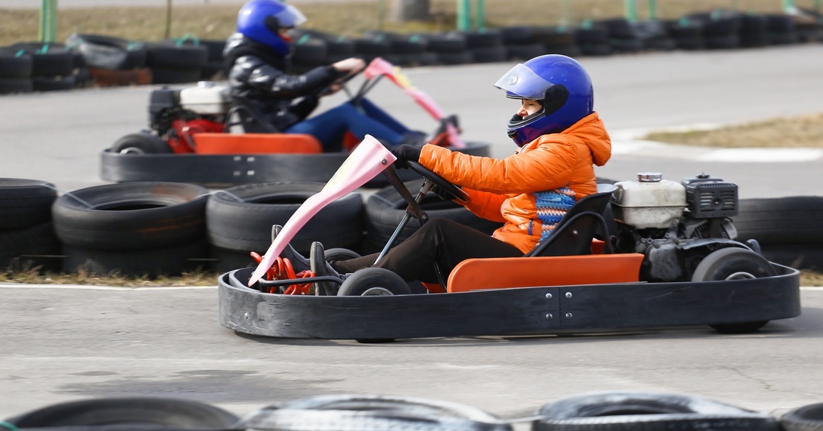 Two racers wearing helmets and protective gear driving go-karts around a track lined with black tires.
