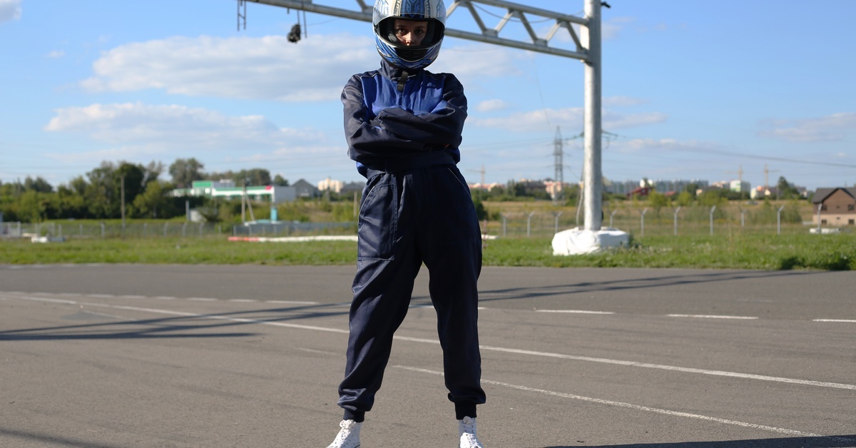 A person wearing a helmet and navy blue racing suit standing with crossed arms on an outdoor go-kart track under clear skies.