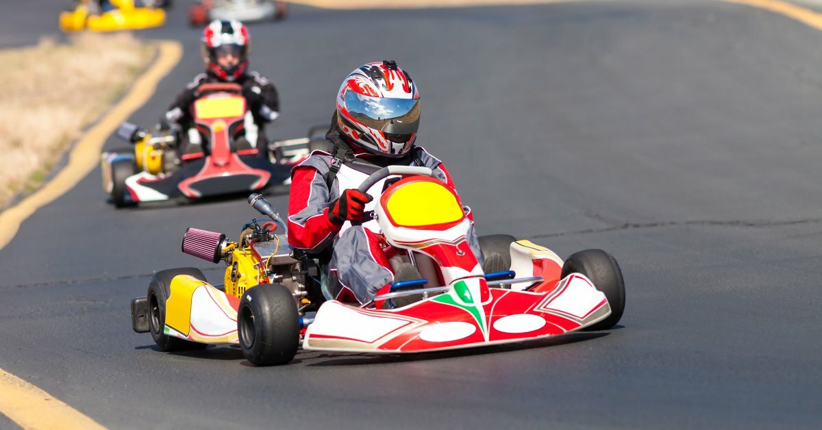 A person wearing a red racing suit and helmet actively driving a go-kart down a paved outdoor track.