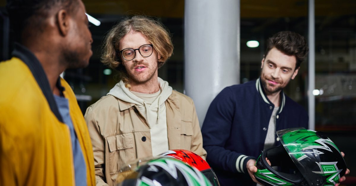 Three people holding brightly colored motorcycle helmets while standing near a go-kart track at night.