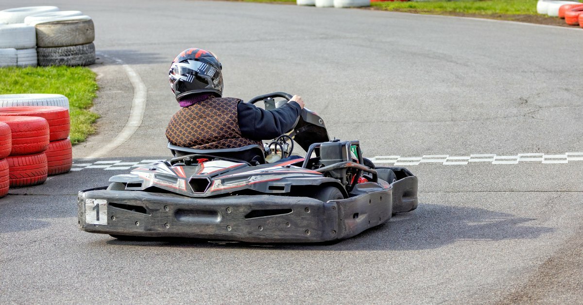A person in a dark vest driving a black go-kart across a checkered finish line surrounded by stacked tire barriers.