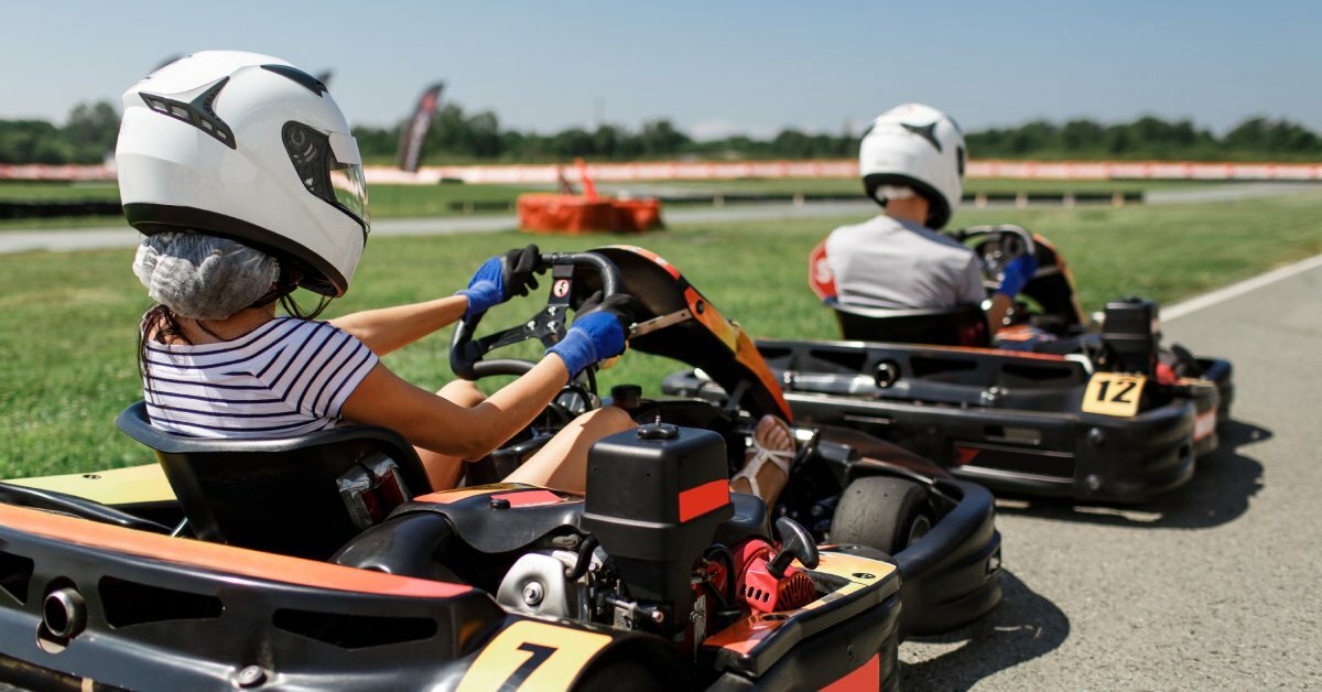Two drivers sitting in numbered go-karts wearing white helmets and gloves near a turn on an outdoor track.