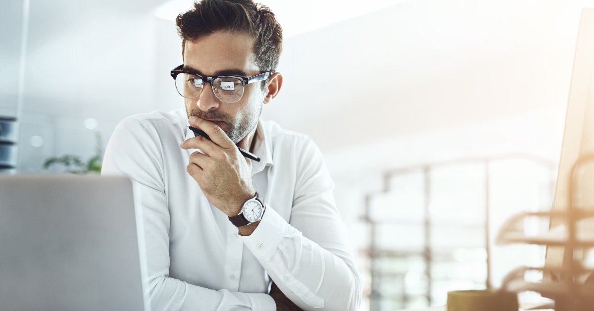 A businessman with his hand on his chin, looking at his laptop. He has a pen in that hand and a notebook in front of him.