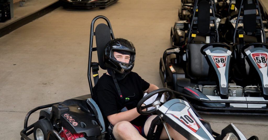 A young man in a helmet and black shirt sitting in a go-kart parked near a row of other go-karts in a garage area.