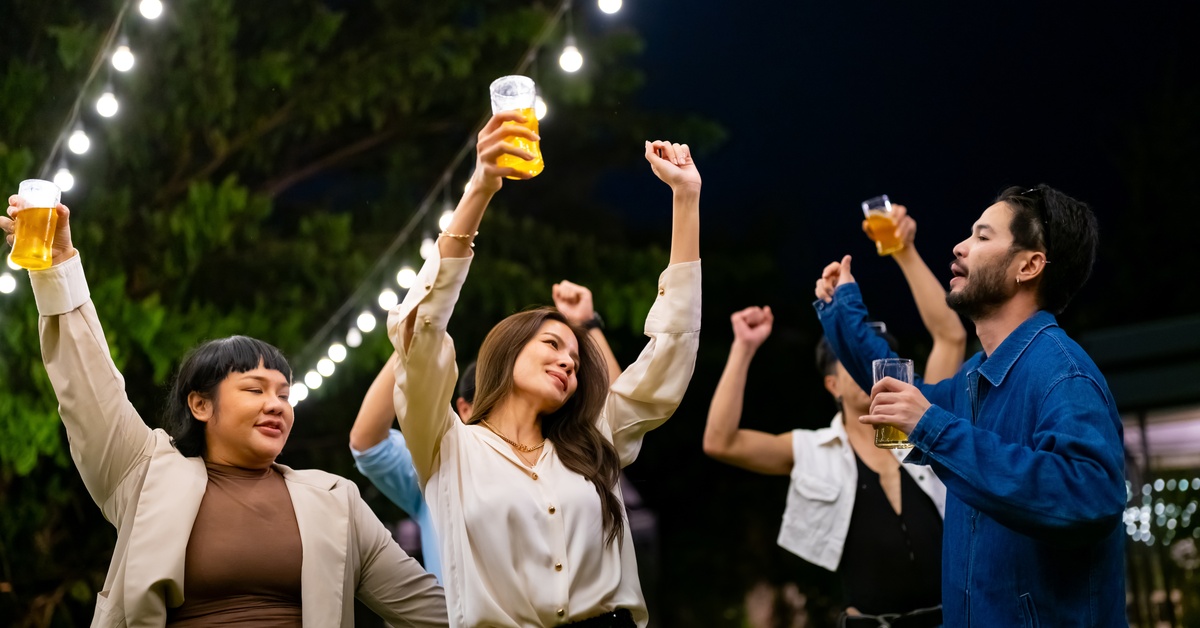 A group of people holding drinks raise their arms and dance under string lights at night in an outdoor setting