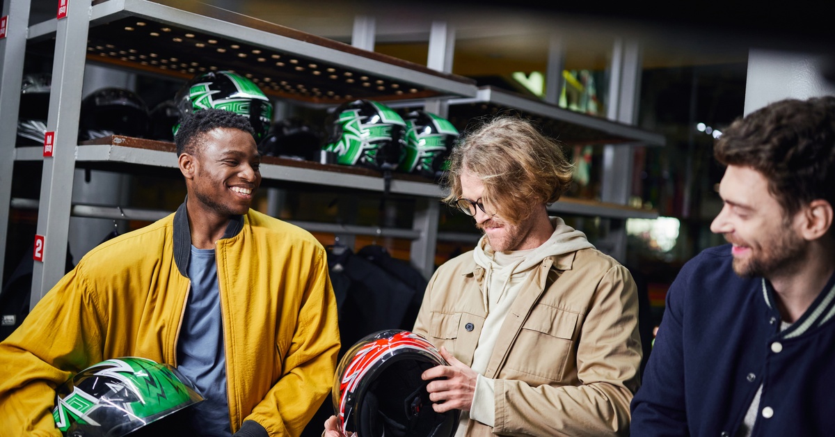 Three men smile and inspect motorcycle helmets in front of a rack filled with green and black helmets.