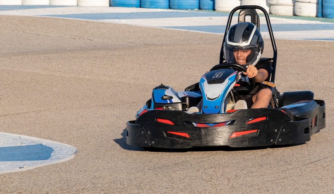 A young racer wearing a helmet drives a blue go-kart on a paved track bordered by colorful tire barriers.