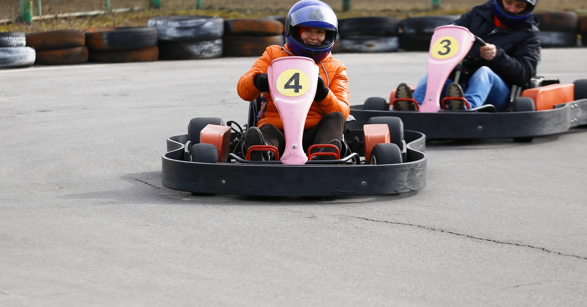 Two people in helmets racing pink go-karts on a paved course with black tire barriers and green fencing.