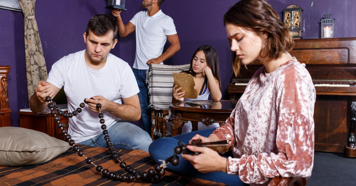 Two people sitting and two standing in a room with dark walls and vintage furniture examine objects and paper clues.