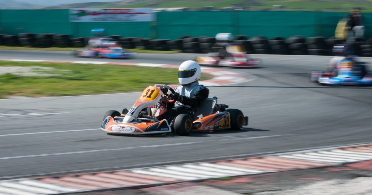 A go-kart driver wearing a white helmet and black suit racing on an outdoor track with other karts visible in the background.