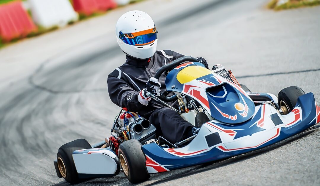 A go-kart racer in a white helmet and black suit speeding down an outdoor track lined with red and white safety barriers.