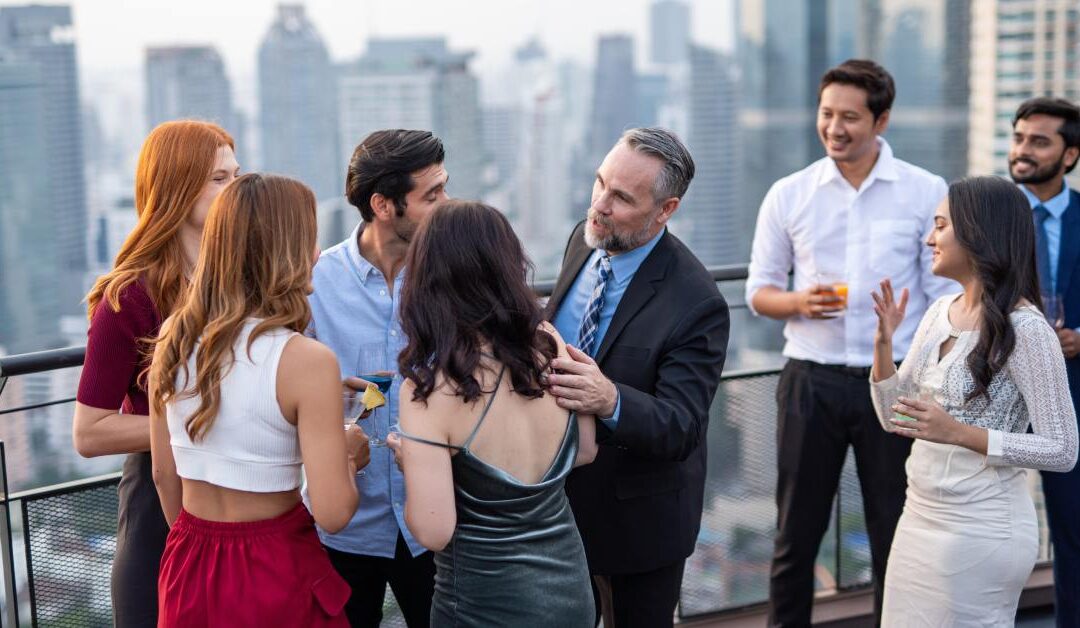 Group of people in formal and semi-formal attire socializing on a rooftop terrace with a city skyline in the background.