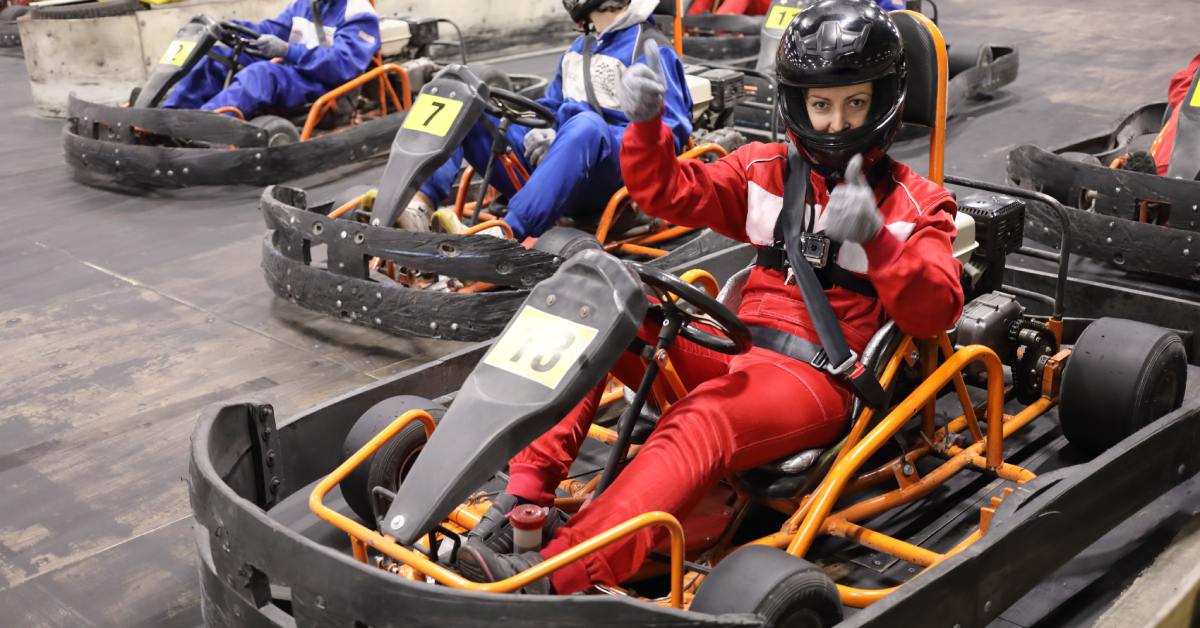 An indoor go-kart track with drivers wearing helmets and racing suits seated in go-karts ready to compete.