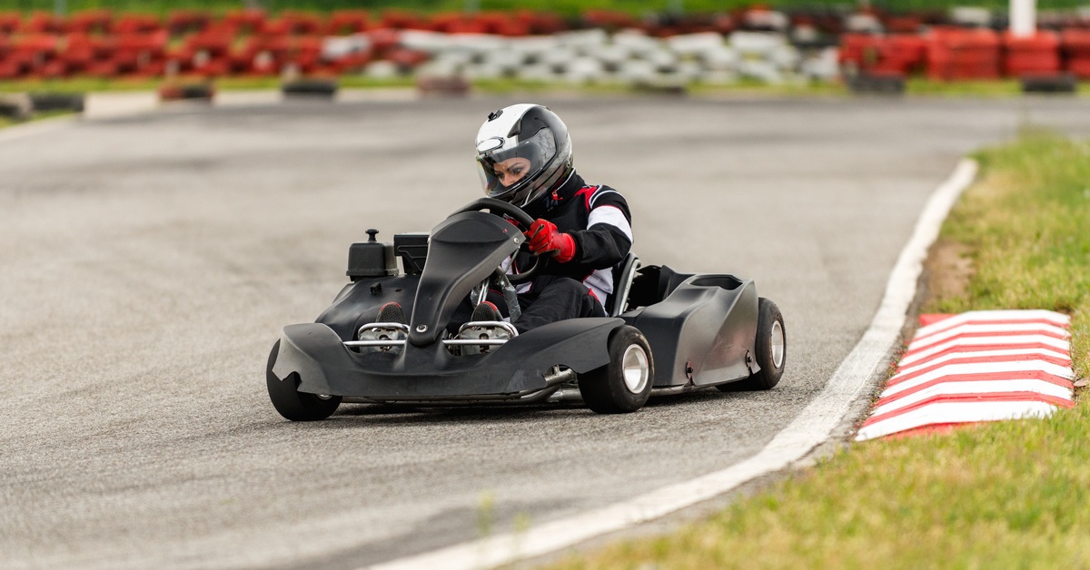 A go-kart driver wearing a helmet and gloves races at high speeds on an outdoor track while approaching a corner.