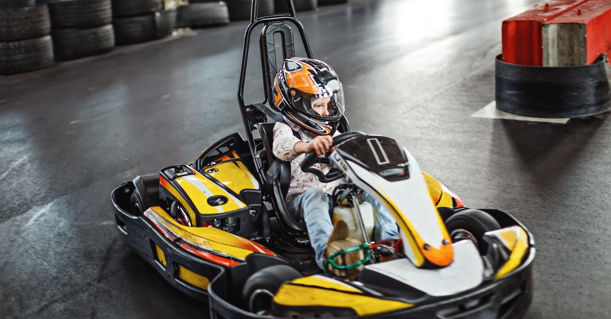 A kid wearing jeans and a floral shirt wears a yellow helmet while driving a yellow and black go-kart around a turn.