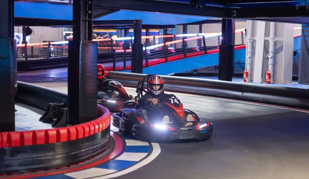 Two people wearing dark clothing and red helmets are driving red and black go-karts on an indoor track.