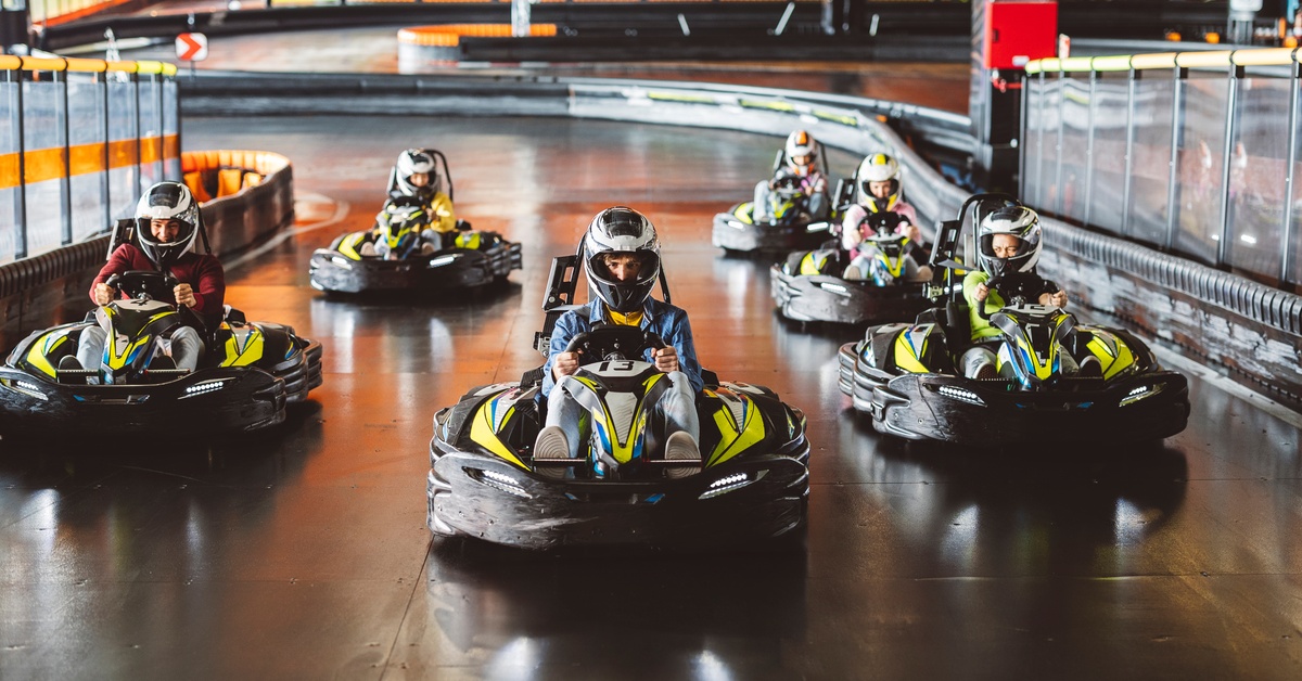 A group of six people wearing black and silver helmets are driving yellow and black go-karts on an indoor track.