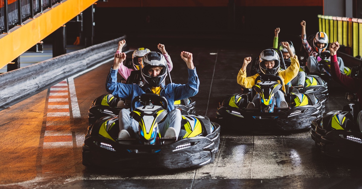 Six people wearing helmets lift their arms into the air while sitting in go-karts on an indoor track.