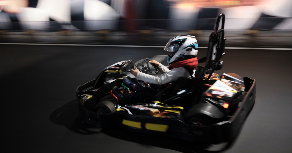 A person wearing a white and teal helmet is driving a black and yellow go-kart on an indoor track.