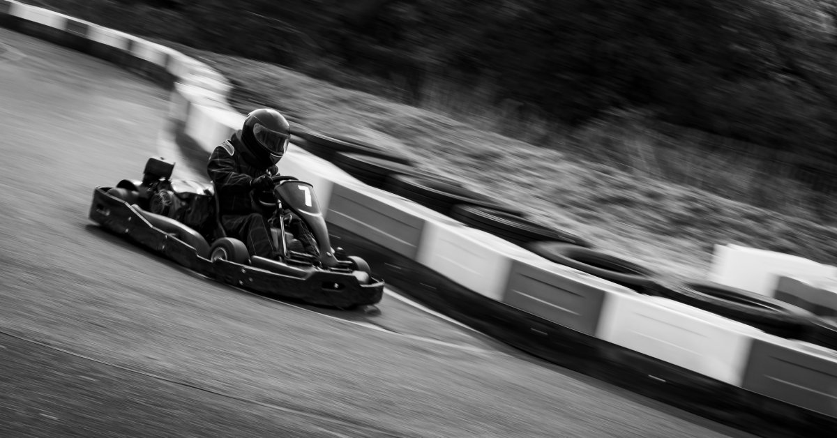A black-and-white view shows a person racing around a curve on a go-kart track. Trees are blurry in the back.