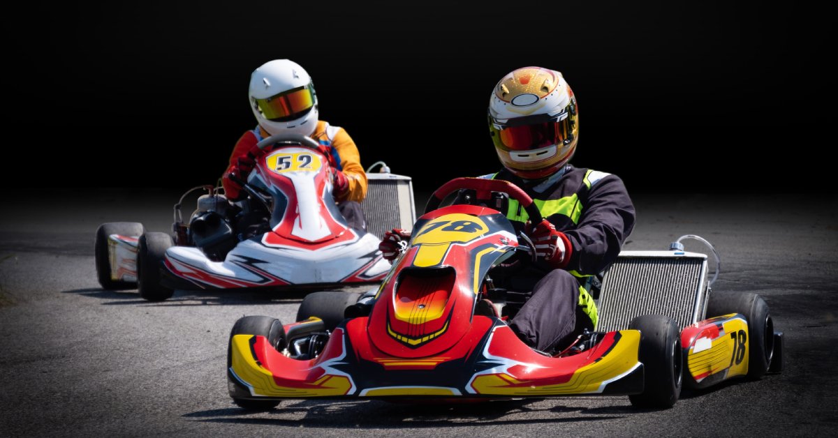Two racers sit in a go-kart on a track. One is wearing a gold helmet and the other is wearing a white helmet.