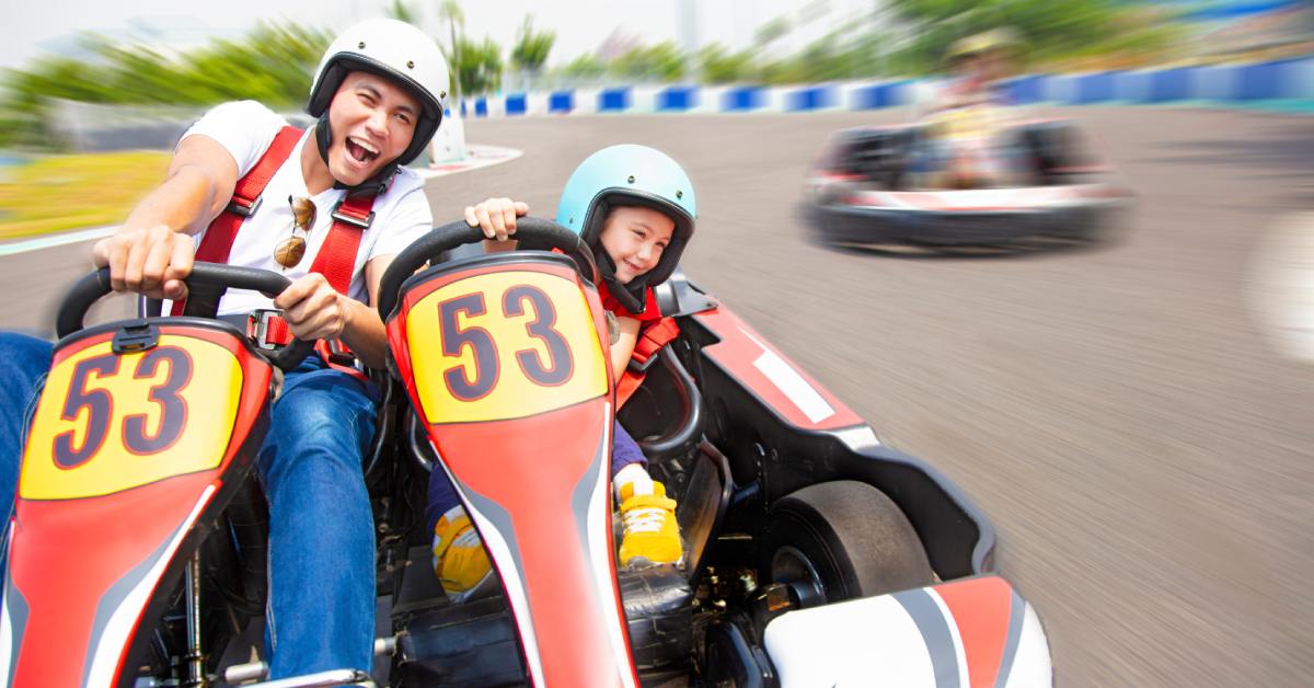 A father and his young daughter are smiling as they drive red and white go-karts on an outdoor track.