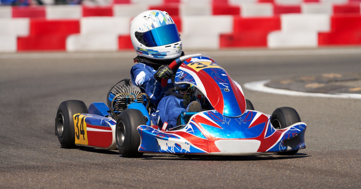 A person wearing a white helmet is driving a blue and red go-kart on an outdoor track. Two people appear blurry behind them.