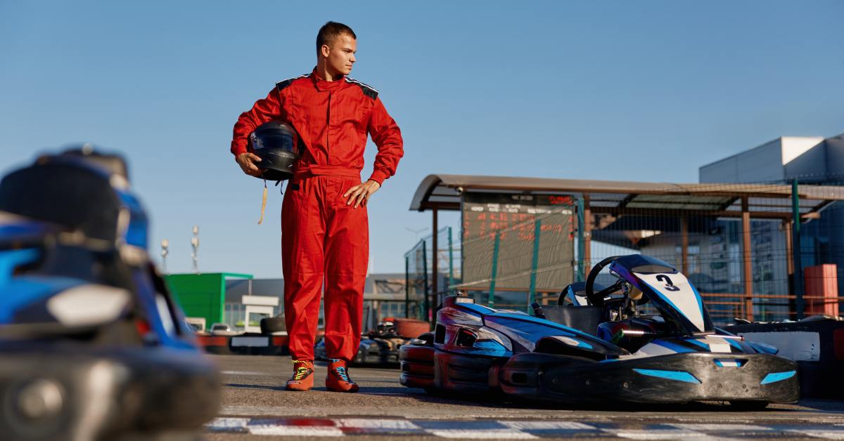 A man wearing a red jumpsuit holds a black helmet. He's standing next to a go-kart at an outdoor track.