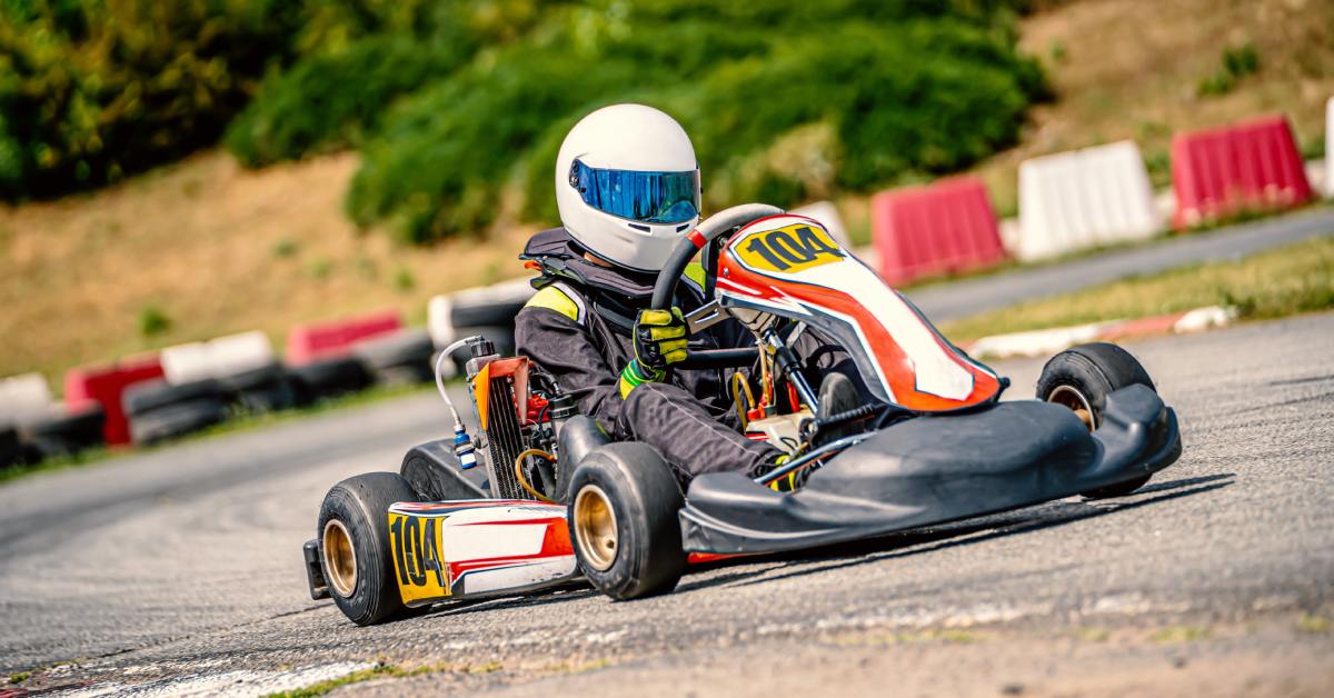 A person is driving a red, black, and white go-kart on an outdoor track. They are wearing a white helmet.