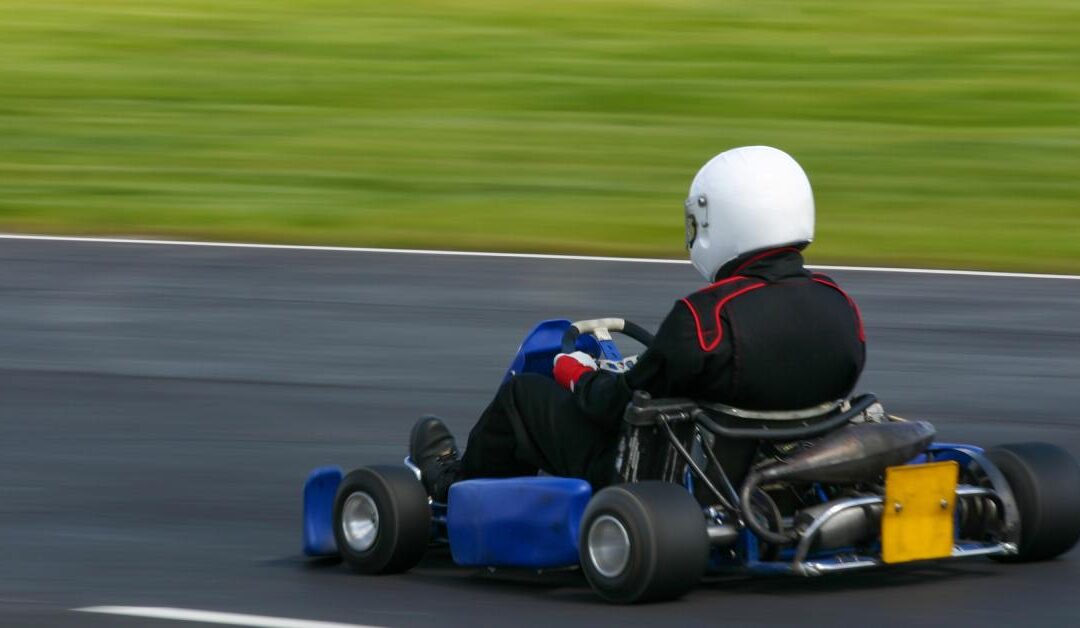 A person wearing a black bodysuit and a white helmet, driving a blue go-kart on an outdoor track. The surroundings are blurry.
