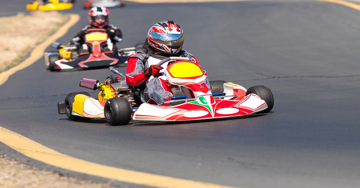 Two people are driving go-karts on an outdoor track. They are going around a curve and wearing helmets.