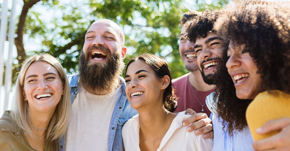 A group of three men and three women smile as they embrace each other. Trees appear behind them.