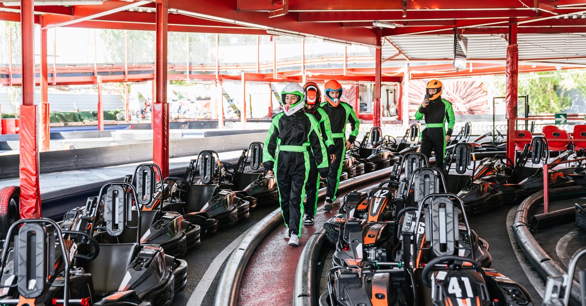 Four people wearing black jumpsuits with neon green stripes down the side are walking between a row of go-karts.