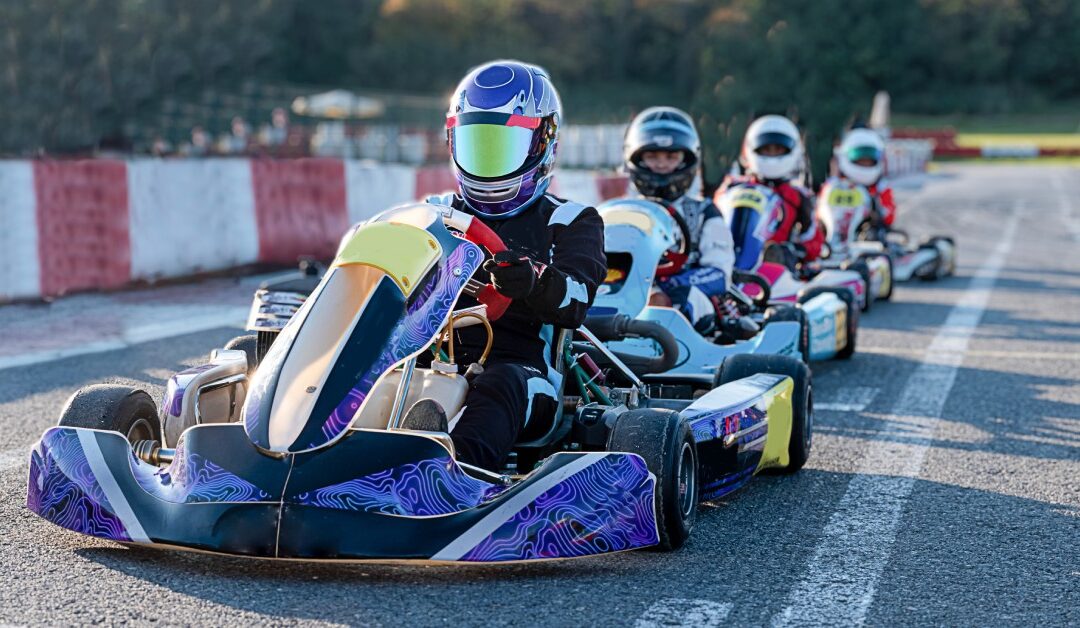 Four people sit lined up behind each other in go-karts on an outdoor track, wearing helmets. The background is blurry.