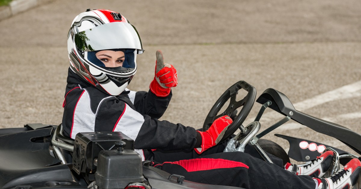 A person wearing a go-kart suit and a white helmet sits in a go-kart on an outdoor track is giving a thumbs-up.