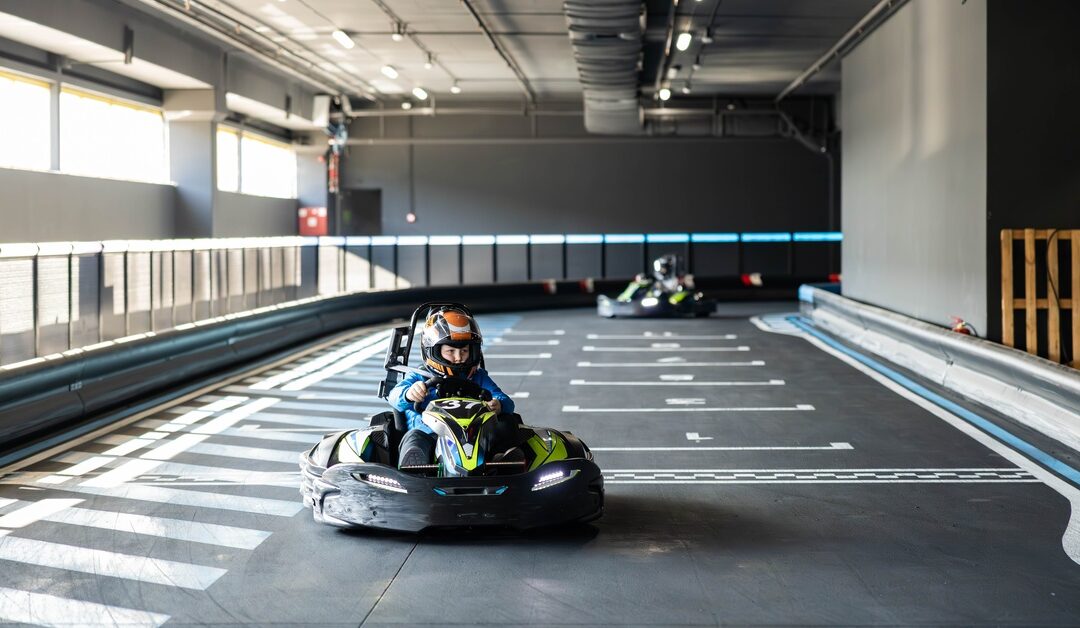 A person is driving a black and bright yellow go-kart wearing a black and orange helmet. Another driver is behind them.