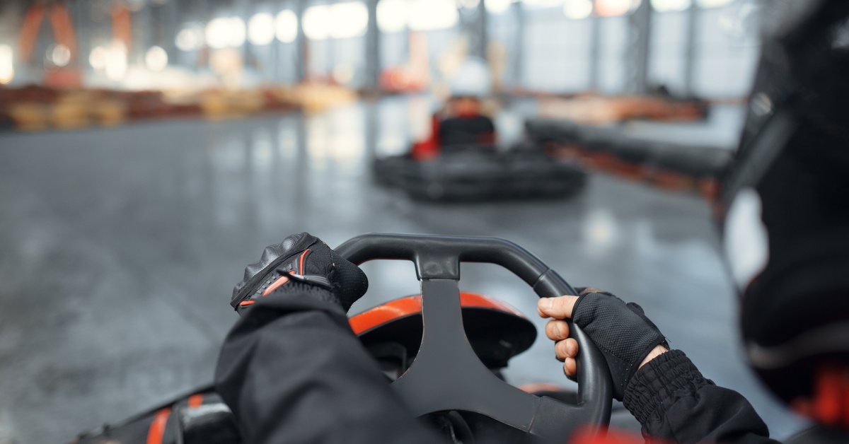 An over-the-shoulder view shows a person holding onto a black steering wheel of a go-kart while wearing gloves.