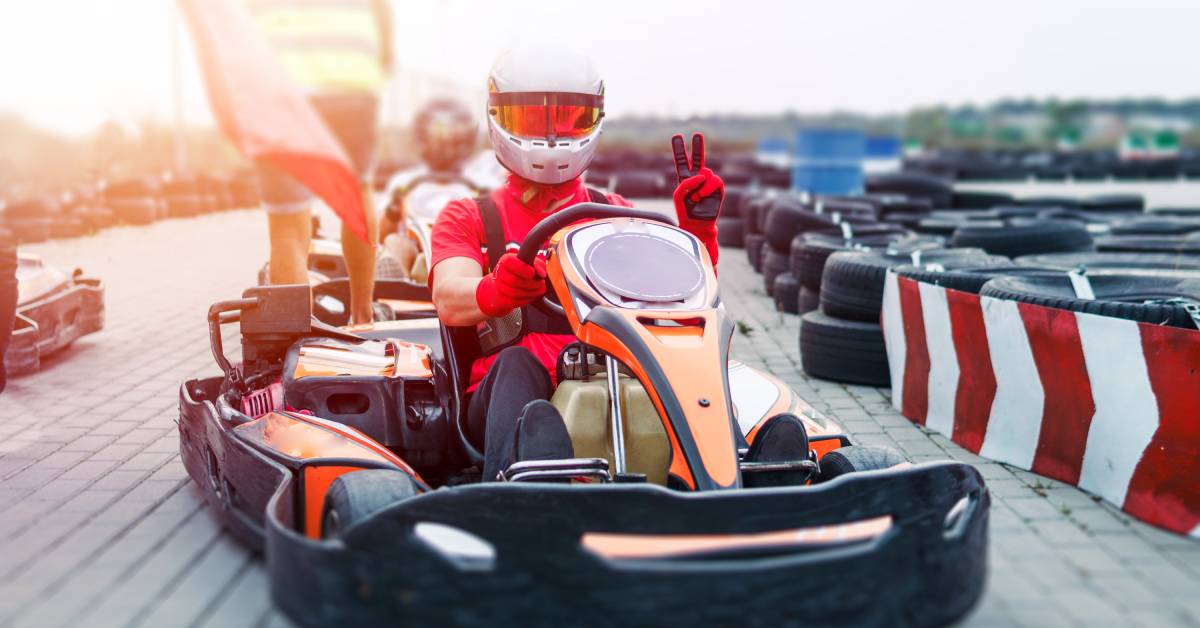 A person wearing a red shirt, a white helmet, and red driving gloves is giving the peace sign while sitting in a go-kart.