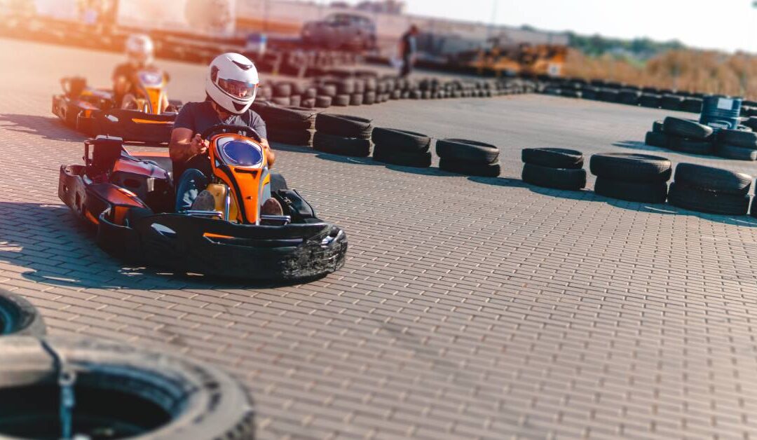 Two people are driving orange and black go-karts on an outdoor track. They're wearing white helmets.