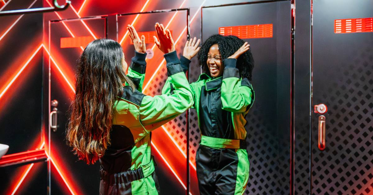 Two women wearing black and bright green jumpsuits are smiling and high-fiving each other. They are in a locker room.
