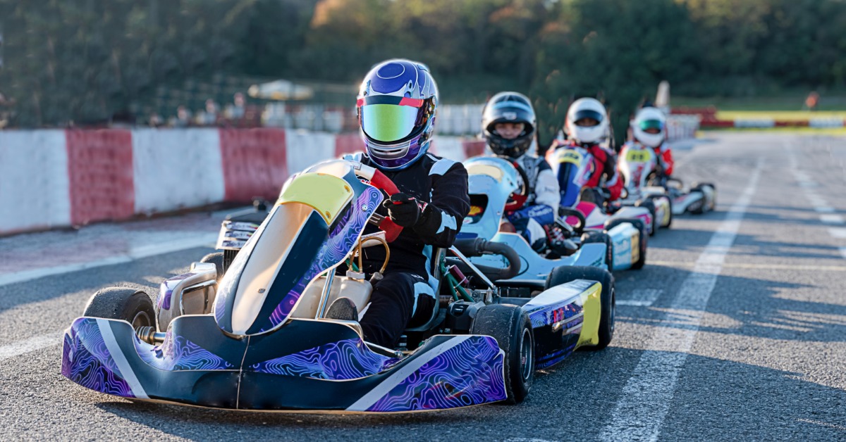 Four go-kart drivers wearing protective helmets are sitting in go-karts that are in a line on an outdoor track.