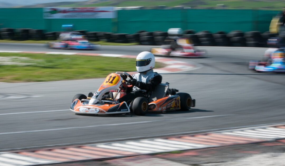 A person wearing a white helmet sits in an orange go-kart on an outside track. Three other go-karts are blurry behind it.