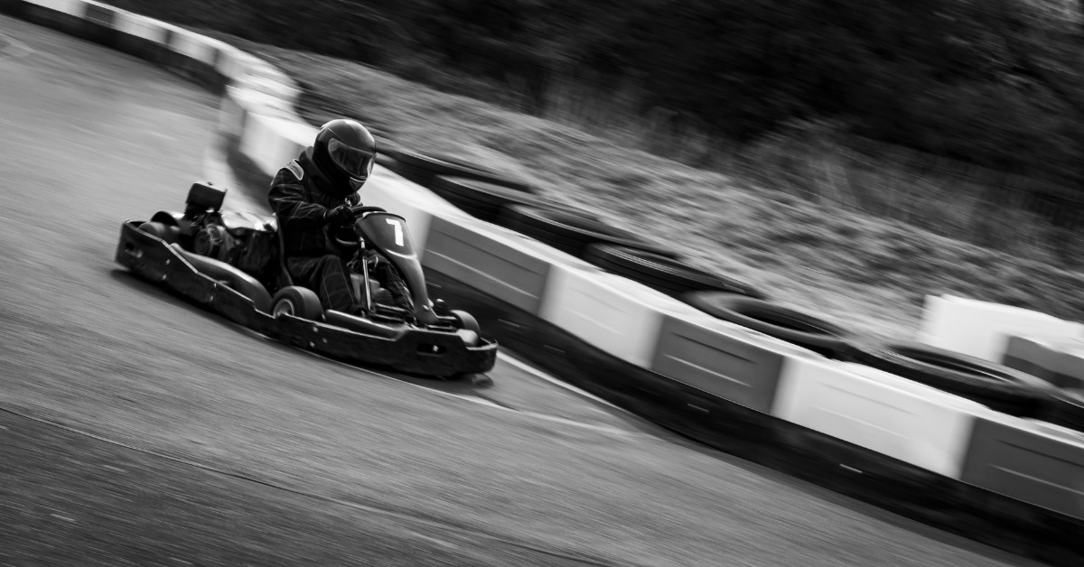 A black and white photo shows a person driving a go-kart quickly on an outdoor track. The person is wearing a helmet.