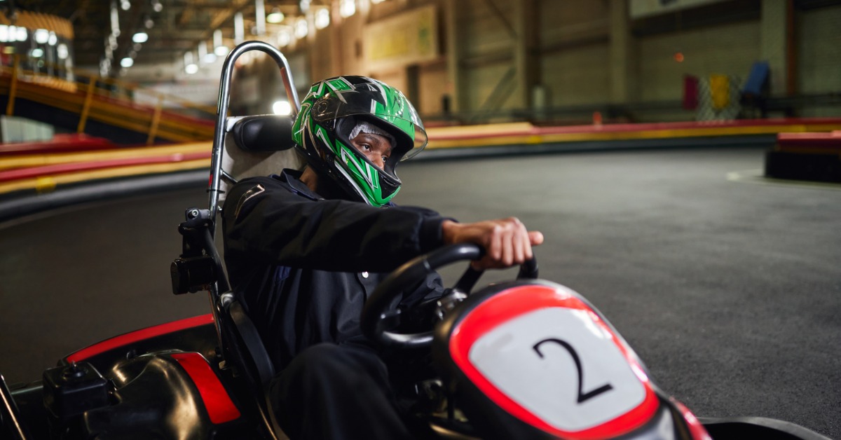 A man wearing a black long-sleeve shirt and a green helmet is driving a red and black go-kart on an indoor track.