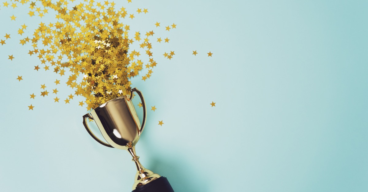 A gold cup with a black base is lying on its side with gold confetti stars emerging from the cup.
