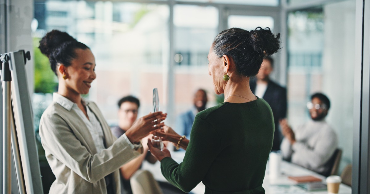 A woman is handing another woman an award while they stand in a conference room with other people.
