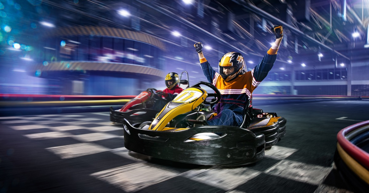 Two people are driving go-karts across the finish line on an indoor go-kart track. One person is raising their arms in the air.