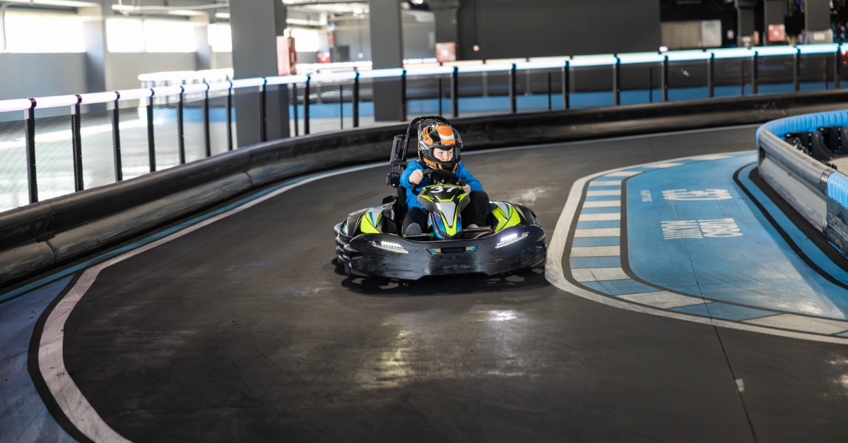 An older child is driving a neon-yellow and black go-kart around a curve on an indoor track. The child is wearing a helmet.