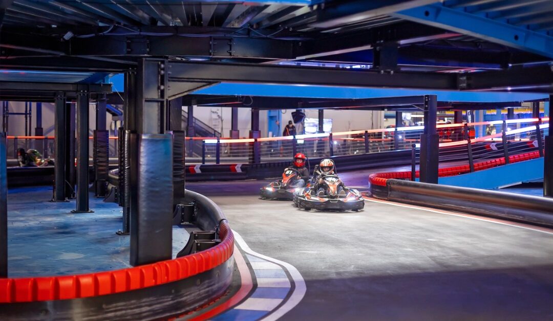 Two people are driving go-karts around the curve on an indoor go-kart track. They are wearing helmets.